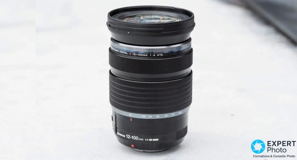 M.Zuiko 12-100mm f/4 IS PRO, vue frontale debout sur fond gris, échelle focales 12-100 mm et inscription 1:4 ø72 visibles