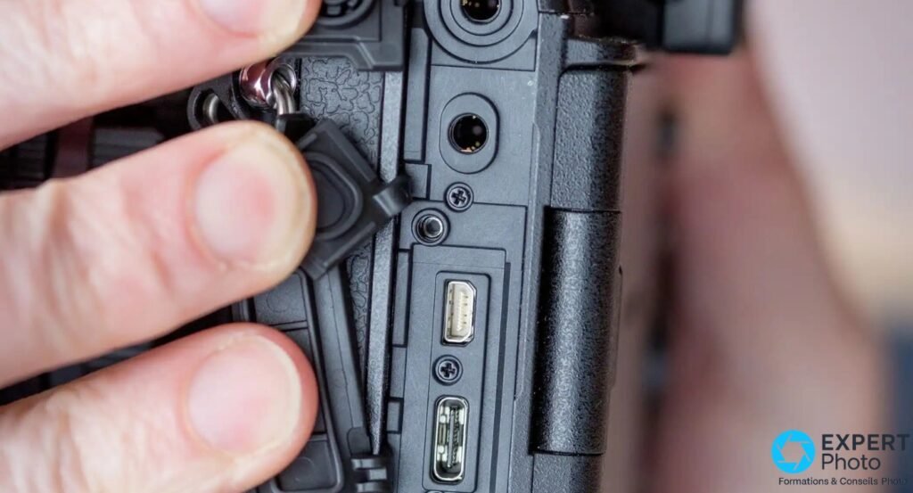 Gros plan sur la trappe de connectiques du flanc gauche de l'Olympus E-M1X : prise jack 3,5 mm et port USB-C visibles, main tenant la trappe