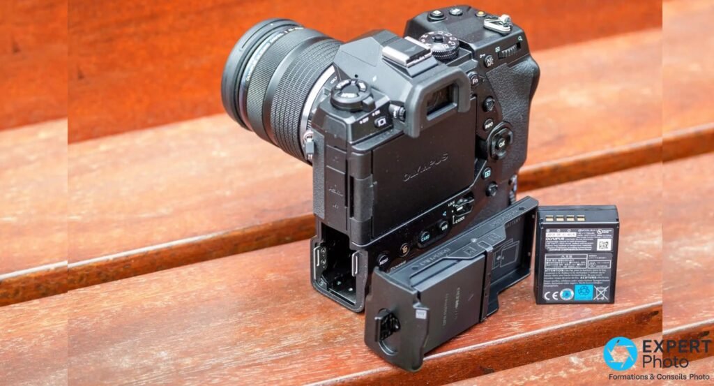  Olympus OM-D E-M1X de dos, trappe du double compartiment batterie ouverte, une batterie Olympus extraite posée sur la surface en bois
