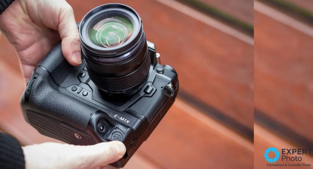 Olympus OM-D E-M1X vu du dessus, tenu à deux mains, inscription E-M1X lisible sur le grip intégré, objectif zoom monté