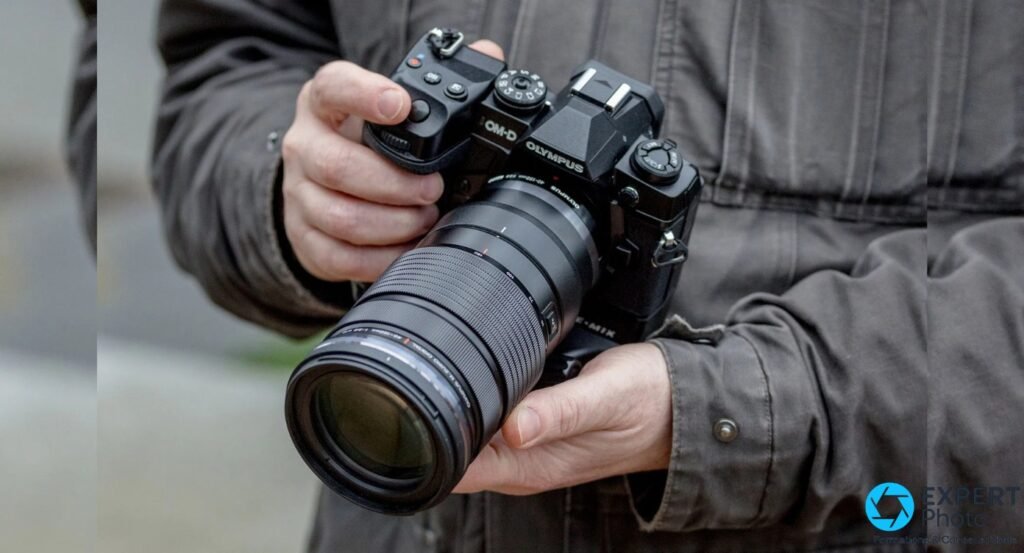 Olympus OM-D E-M1X tenu à deux mains en extérieur, équipé d'un long téléobjectif zoom noir, fond urbain flou
