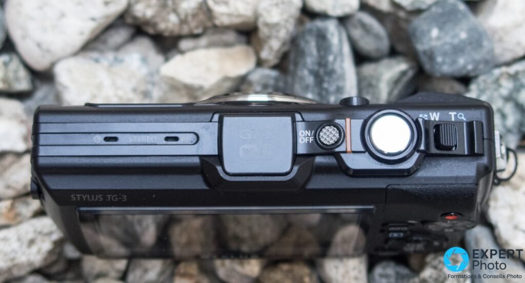Dessus de l'Olympus Tough TG-3, bouton ON/OFF, déclencheur, molette de zoom et sabot GPS/Wi-Fi visibles, fond de galets