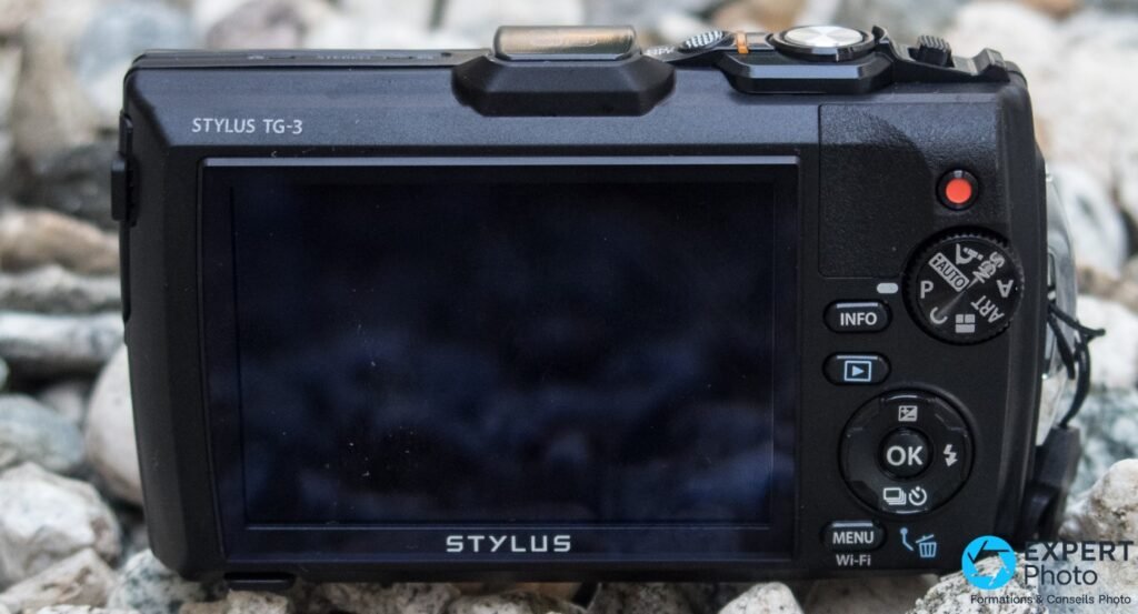 Dos de l'Olympus Tough TG-3, grand écran LCD éteint, molette de modes AUTO/ART/P visible et bouton vidéo rouge, fond de galets