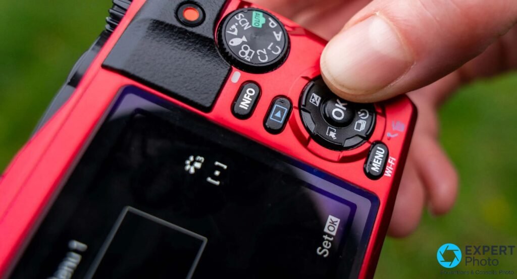 Dos Olympus Tough TG-6 rouge, molette modes AUTO visible, boutons INFO MENU Wi-Fi OK, doigt sur la molette, fond vert flou
