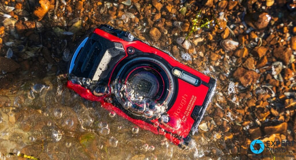 Olympus Tough TG-6 rouge mouillé posé dans l'eau sur fond de galets, objectif vers le haut, eau et bulles visibles