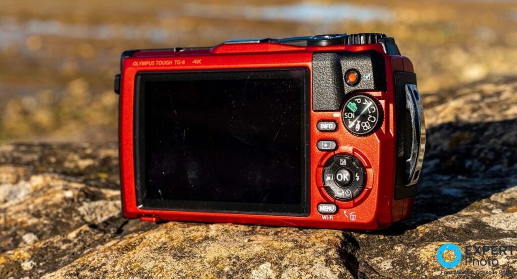 Dos Olympus Tough TG-6 rouge sur roche, écran LCD éteint, mention TOUGH TG-6 4K, molette modes et boutons MENU Wi-Fi