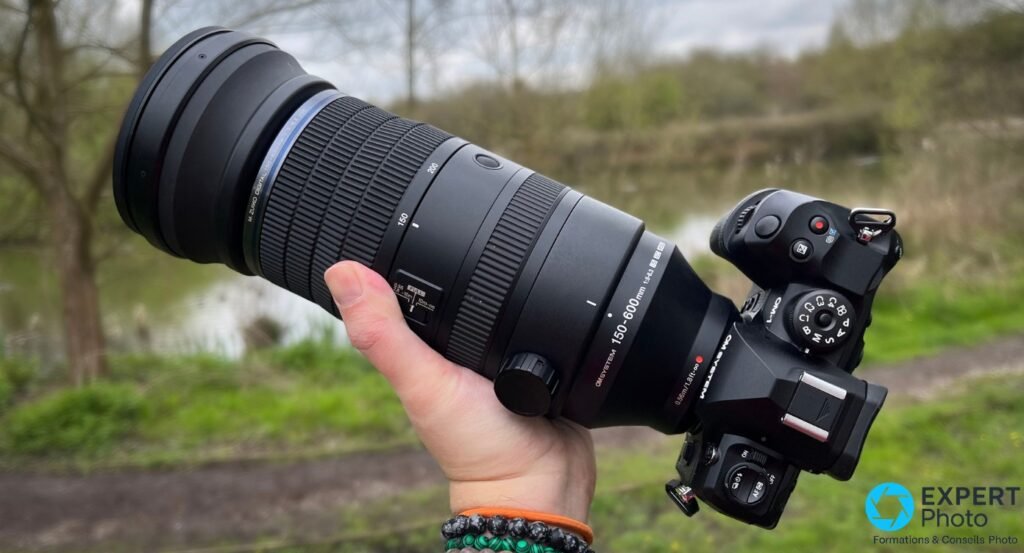 OM System M.Zuiko 150-600mm monté sur boîtier OM System, tenu de profil, fond bord d'eau et arbres dénudés 