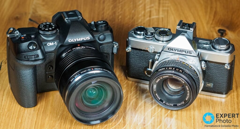 OM System OM-1 avec M.Zuiko 12-40mm et ancien boîtier Olympus argentique Zuiko Auto-S 50mm, posés sur bois