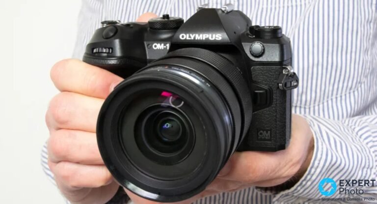 OM System OM-1 tenu à deux mains face à l'objectif, marquage OM-1 et Olympus lisibles, objectif zoom monté