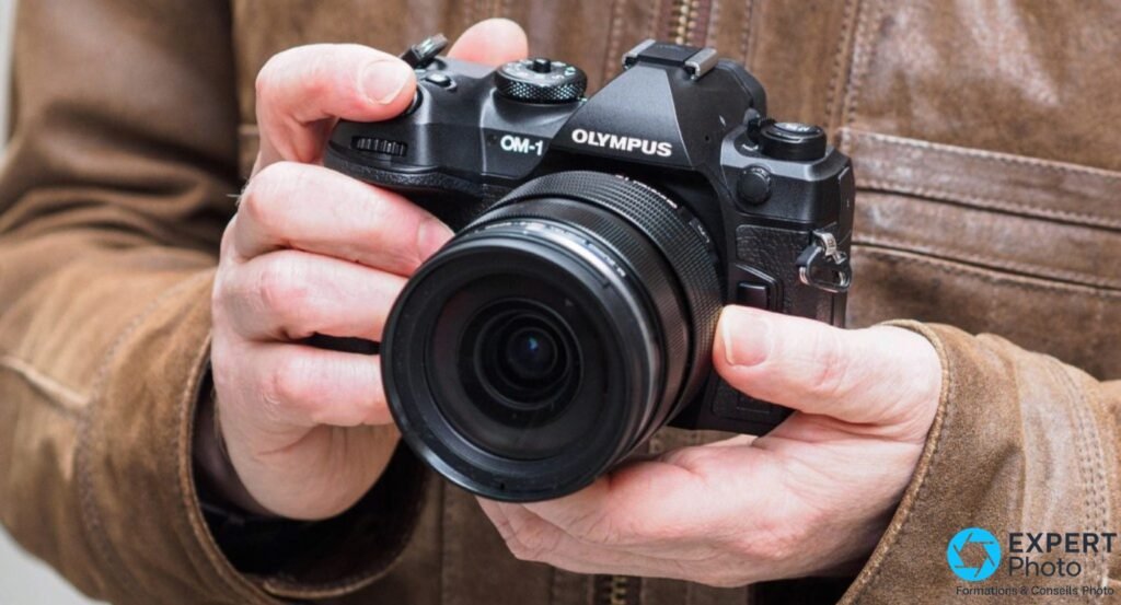 OM System OM-1 tenu à deux mains, vue de face légèrement inclinée, marquage OM-1 et Olympus lisibles, veste marron