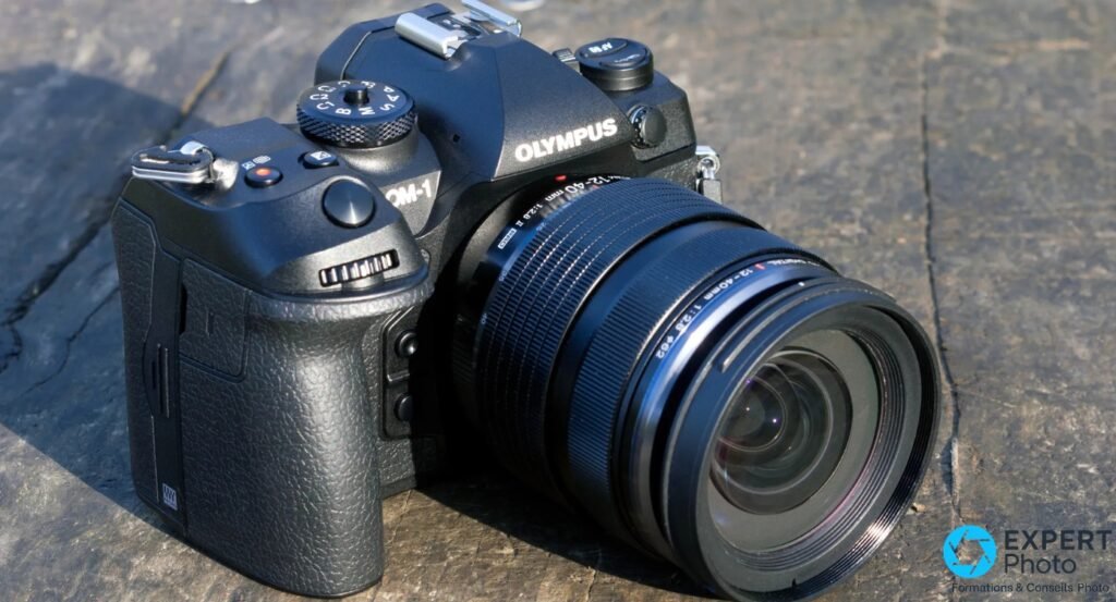 OM System OM-1 posé sur pierre, vue 3/4 avant droit côté grip, objectif M.Zuiko 12-40mm f/2.8 II, marquage PRO lisible 