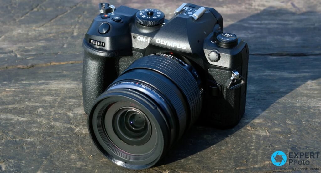 OM System OM-1 posé sur pierre, vue 3/4 avant gauche, marquage M.Zuiko Digital 12-40mm lisible sur l'objectif 