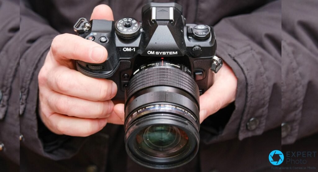  OM System OM-1 Mark II noir tenu à deux mains, objectif M.Zuiko 12-40mm f/2.8 en avant, prise en extérieur