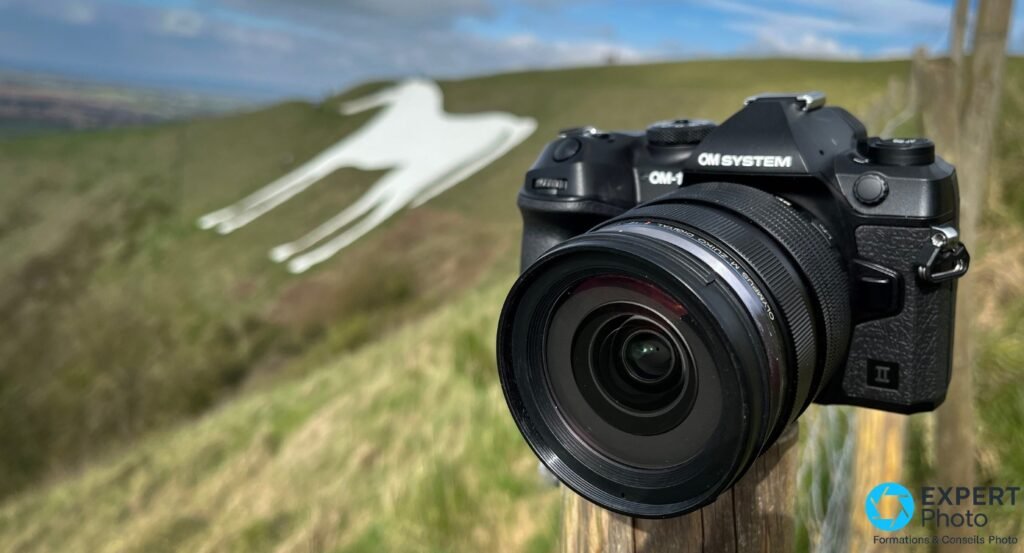  OM System OM-1 Mark II noir avec zoom M.Zuiko, posé sur un pieu en extérieur, colline verte en arrière-plan