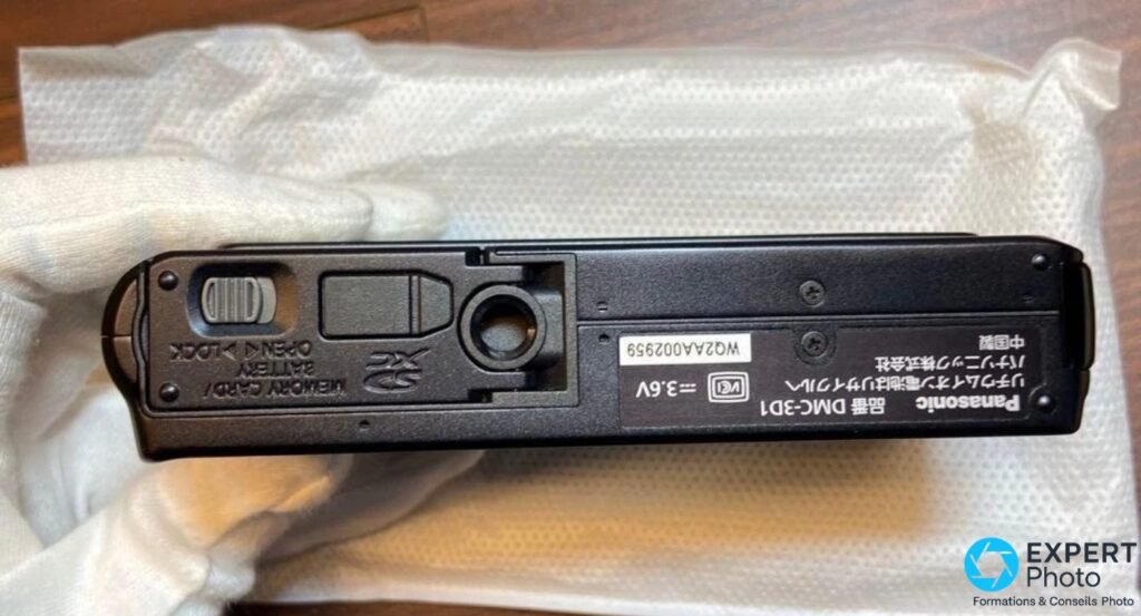Dessous Panasonic DMC-3D1 en mains gantées, trappe batterie-carte mémoire ouverte, compartiment vide visible