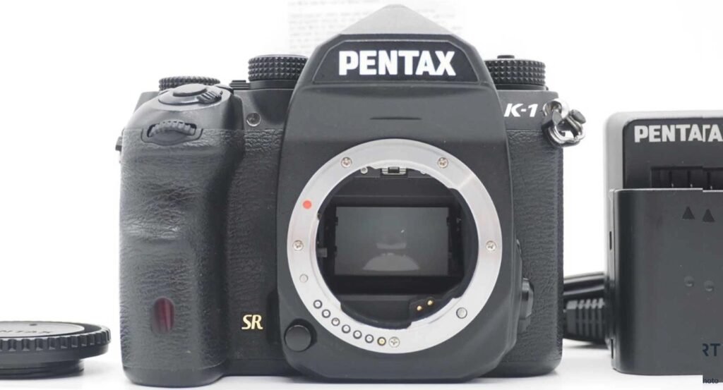 Pentax K-1 noir de face, boîtier nu monture ouverte, chargeur Pentax et cache objectif posés à côté