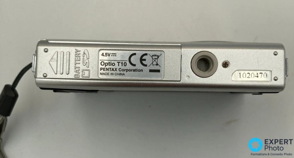 Dessous du Pentax Optio T10 : étiquette blanche avec mention Optio T10 PENTAX Corporation Made in China, numéro 1020470, prise ronde et trappe batterie