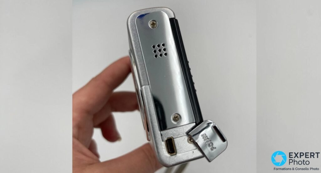  Pentax Optio T10 tenu en main, tranche gauche avec grille de haut-parleur, prise de connexion et clip de dragonne estampillé Pentax
