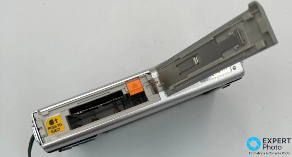 Pentax Optio T10 : trappe inférieure ouverte révélant les logements batterie et carte SD vides, éjecteur orange et étiquette PUSH TO EJECT visibles