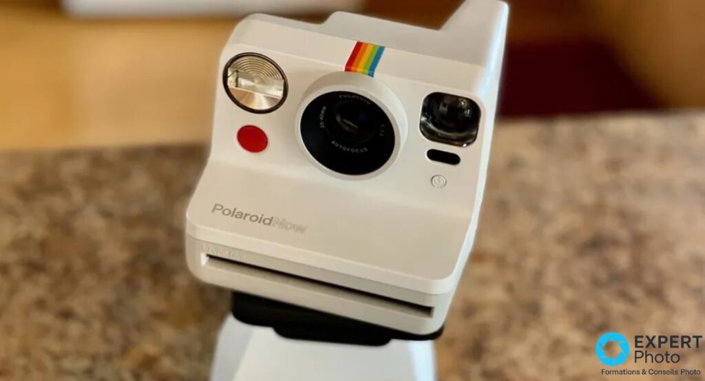 Polaroid Now Gen 1 blanc, face avant en légère contre-plongée, objectif AUTOFOCUS 35-40mm, flash, bouton rouge, bande arc-en-ciel en haut du boîtier