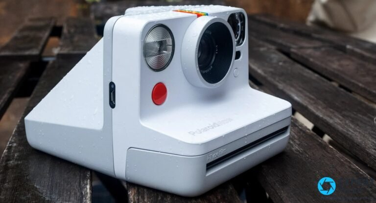 Polaroid Now 1re génération : avis, limites et achat d’occasion Polaroid Now Gen 1 blanc, vue 3/4 avant droit sur fond bois sombre, flash et objectif AUTOFOCUS 35-40mm visibles, gouttes d'eau sur le boîtier