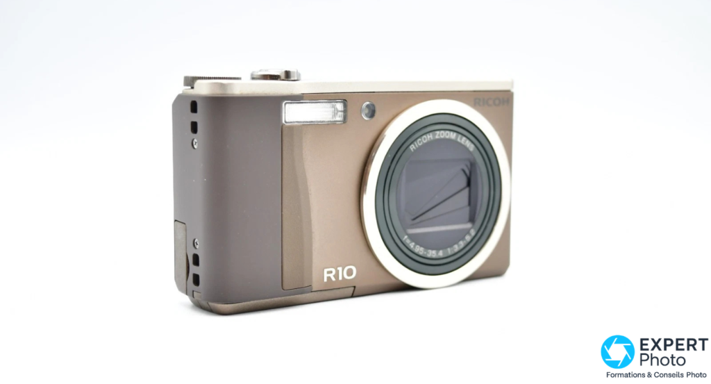 Ricoh R10 en angle 3/4 droit sur fond blanc, zoom rétracté, tranche avec connecteurs latéraux visible, marquage R10 en bas