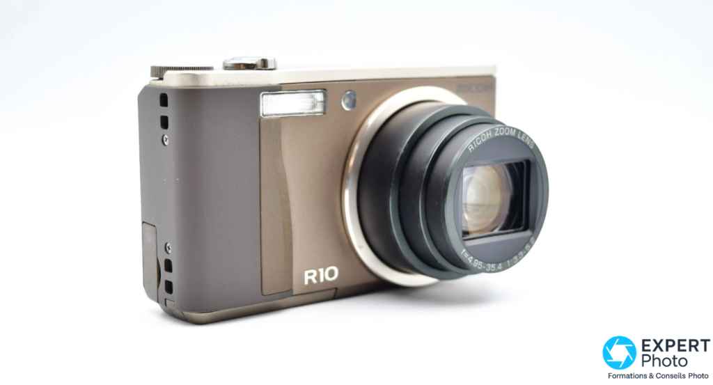 Ricoh R10 en angle 3/4 droit, zoom déployé avec barillet sorti, bague inscrite RICOH ZOOM LENS, fond blanc