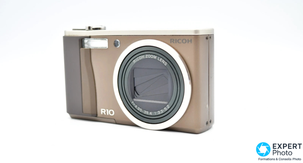 Ricoh R10 de face sur fond blanc, bague objectif inscrite RICOH ZOOM LENS f=4.95-35.4, flash relevé, marquage R10 au bas du boîtier