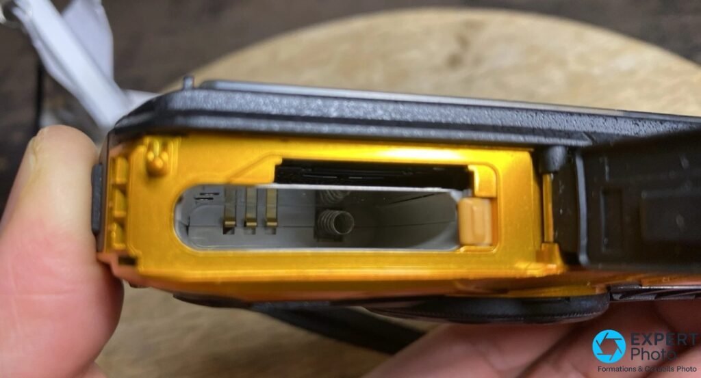 Ricoh WG-30W jaune, compartiment batterie et carte SD ouvert, logements vides, intérieur doré, tenu en main