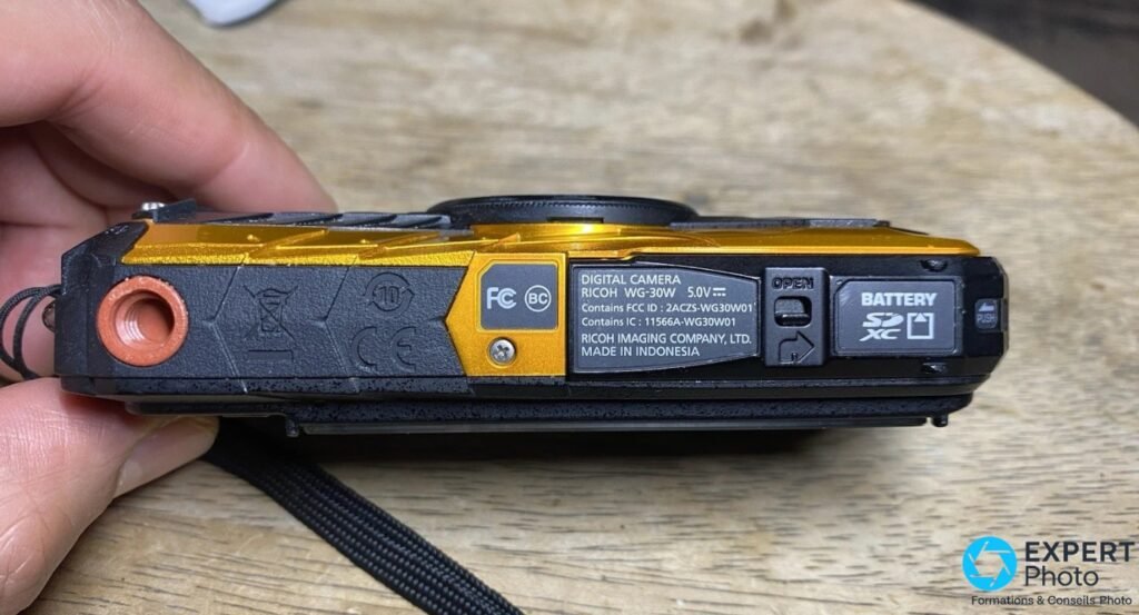 Ricoh WG-30W jaune, tranche inférieure, étiquette RICOH WG-30W lisible, trappes batterie et carte SD, fond bois