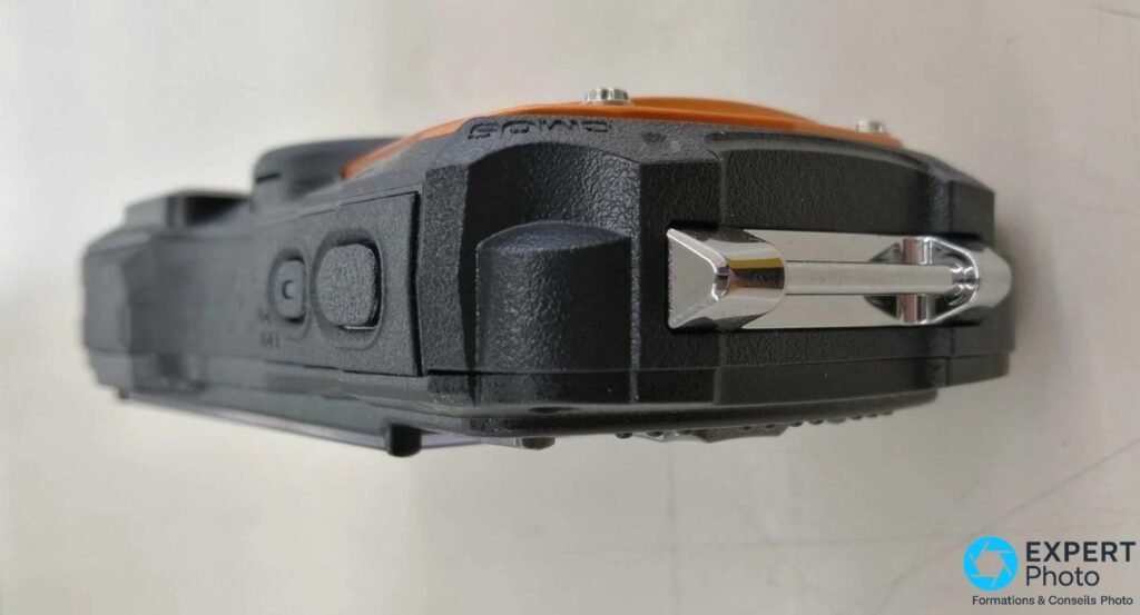 Ricoh WG-80, tranche supérieure, deux boutons noirs et élément métallique argenté en forme de crochet, fond gris neutre