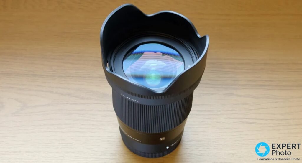Sigma 16mm f/1.4 DC DN Contemporary de face, pare-soleil tulipe monté, lentille frontale visible avec reflets irisés, fond bois clair