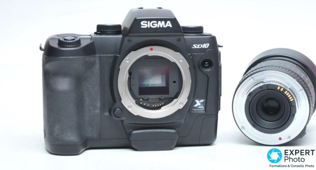 Sigma SD10 de face, monture SA ouverte révélant le capteur Foveon X3 et les contacts électriques, objectif zoom posé à droite