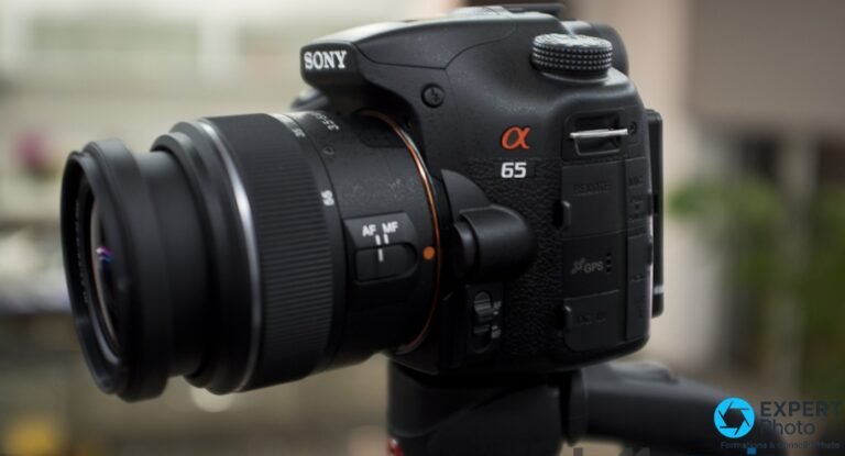 Sony Alpha 65 noir vu de 3/4 gauche avec objectif zoom monté, logo α65 et marquage GPS visibles sur le flanc