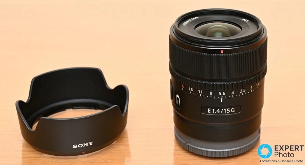 Sony E 15mm f/1.4 G avec pare-soleil Sony noir tulipe posé séparément, inscription E 1.4/15G sur le fût, fond bois clair