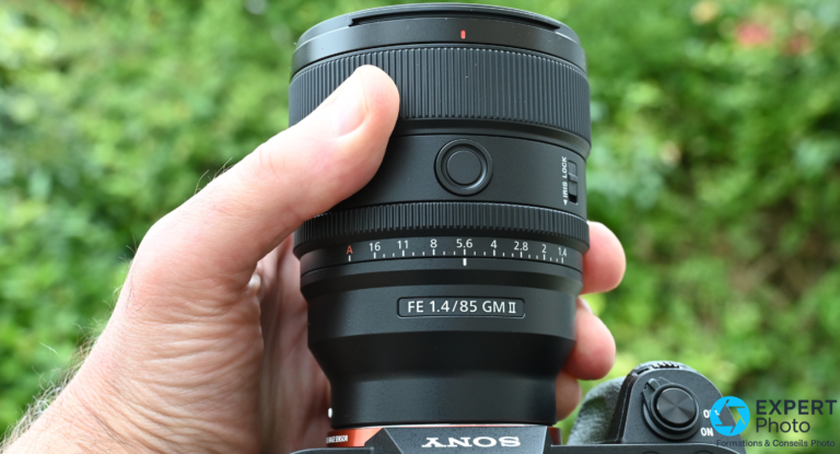 Sony FE 85mm f/1.4 GM II : avis pour portraitistes Sony E Sony FE 85mm f/1.4 GM II en main, inscription FE 1.4/85 GM II, graduation diaphragme et Iris Lock visibles, fond végétation.