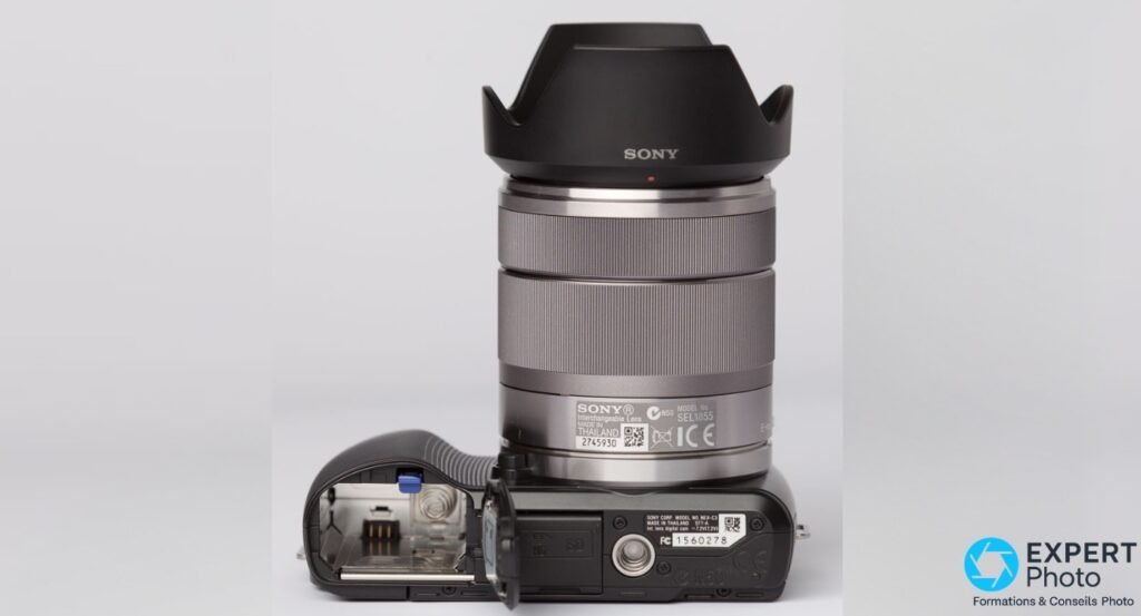 Sony NEX-C3 vu dessous, compartiment batterie et SD ouvert, batterie absente, objectif Sony SEL1855 monté avec pare-soleil.