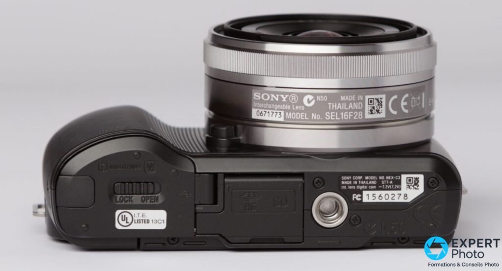 Sony NEX-C3 profil dessous, étiquette modèle NEX-C3 et FC 1560278 lisibles, objectif Sony SEL16F28 monté, trappe SD fermée.