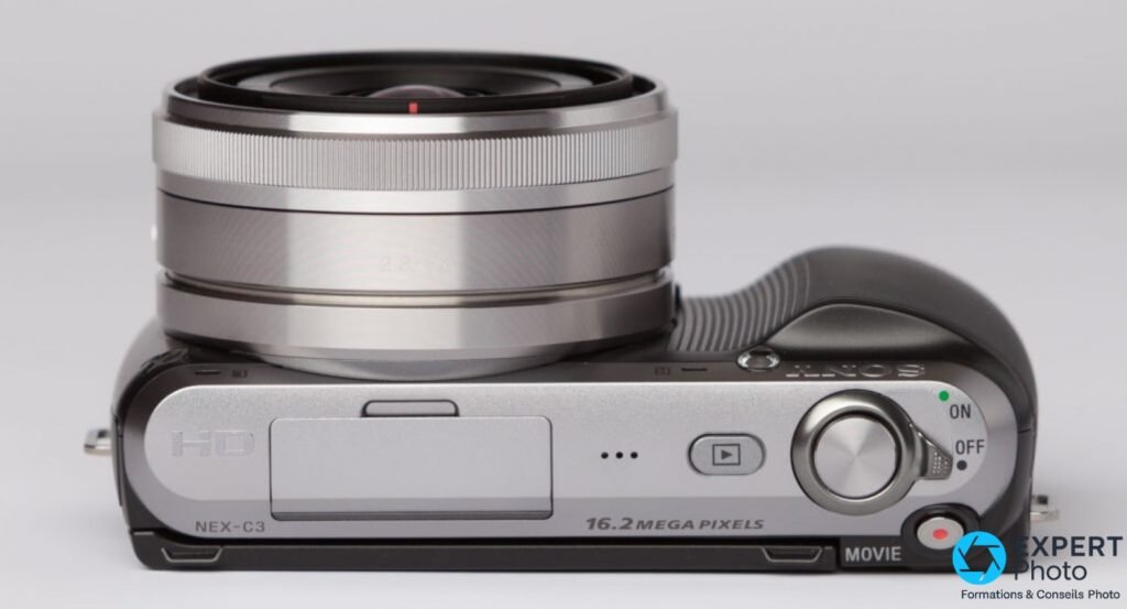 Sony NEX-C3 vue dessus, objectif compact argenté monté, inscriptions NEX-C3 et 16.2 MEGA PIXELS lisibles, fond gris.