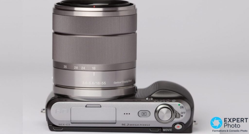 Sony NEX-C3 vue dessus, zoom Sony 18-55 mm f/3.5-5.6 OSS étendu monté, graduation 18-24-28-35 mm lisible, fond gris.