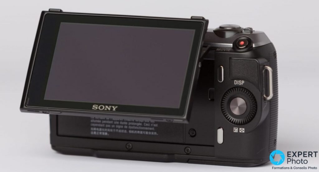 Sony NEX-C3 vue arrière 3/4, écran 3" orientable déplié vers le haut (éteint), bouton MOVIE rouge, molette DISP visibles.