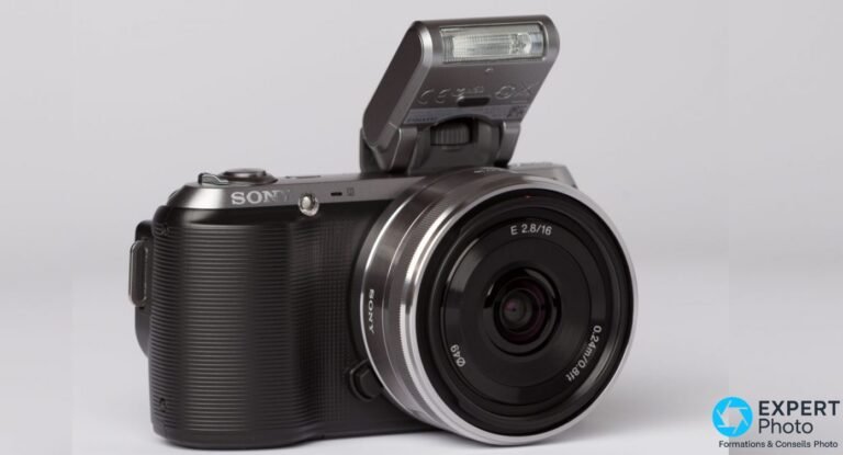 Sony NEX-C3 avis : faut-il encore acheter cet hybride APS-C d’occasion ? Sony NEX-C3 noir avec objectif Sony E 2.8/16 argenté monté, flash amovible déployé, vue 3/4 avant gauche, fond gris neutre.