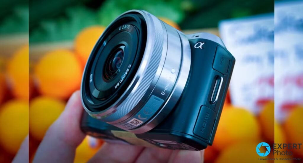 Sony NEX-C3 noir tenu en main, objectif Sony E 2.8/16 argenté monté, fond composé d'oranges et d'un décor de marché.