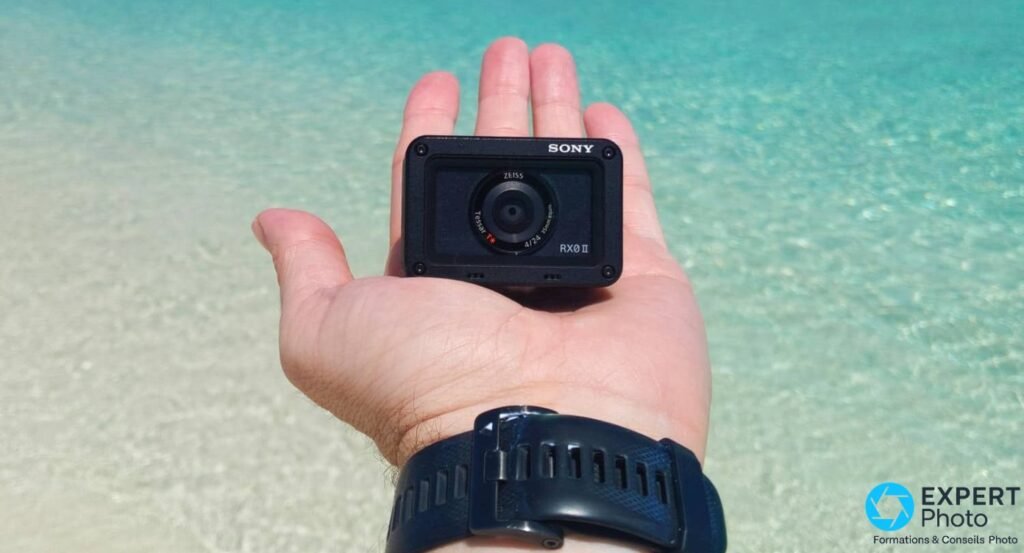 Sony RX0 II noire dans une paume ouverte devant une mer turquoise, compacité illustrée, logo SONY et RX0 II lisibles