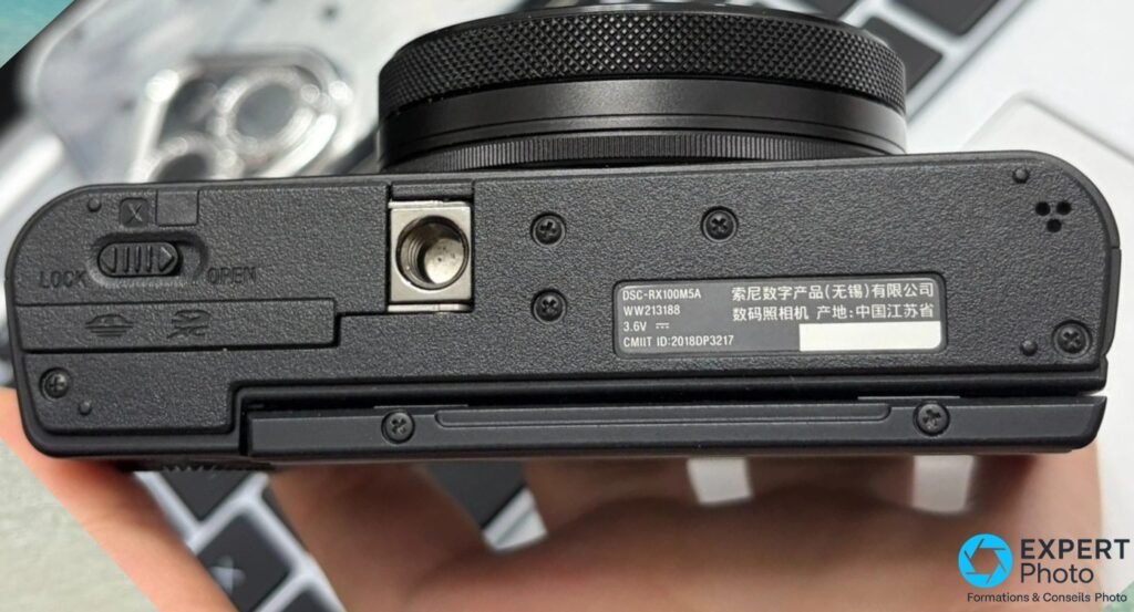 Dessous du Sony RX100 VA avec étiquette DSC-RX100M5A, curseur LOCK/OPEN et filetage trépied, boîtier tenu en main