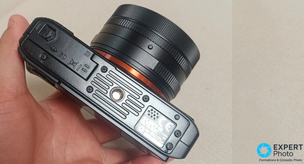 Sony RX1R de dessous : trappe SD et batterie, anneau orange de l'objectif visible, marquage CE sur plaquette, oeillet dragonne gauche 