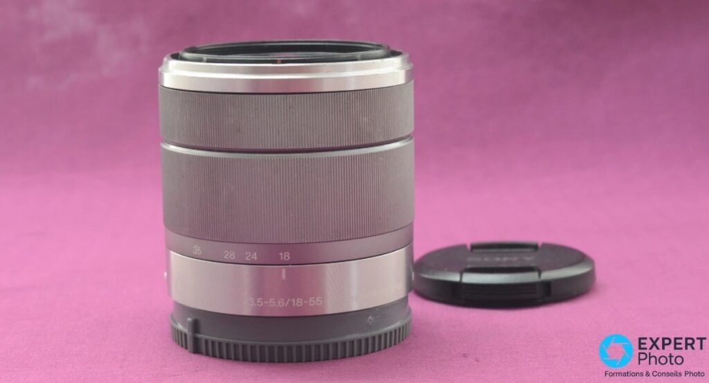 Zoom Sony SEL1855 18-55mm OSS argenté vu de face, bague marquée 3.5-5.6/18-55, cache avant noir Sony posé à droite