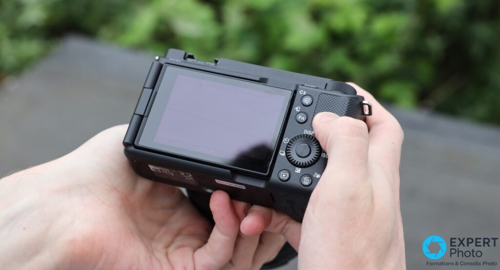 Sony ZV-E1 noir tenu à deux mains, écran orientable déployé, boutons C2, Fn, DISP et molette ISO visibles à l'arrière