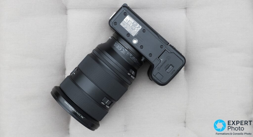 Sony ZV-E1 vu du dessous, trappe batterie avec mécanisme LOCK/OPEN visible, étiquettes réglementaires et bague objectif avec graduations de distance
