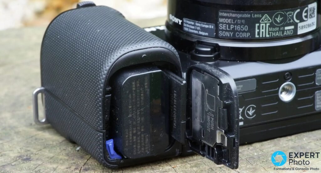 Sony ZV-E10 trappe batterie ouverte, batterie et carte mémoire insérées, objectif Sony SELP1650 en arrière-plan